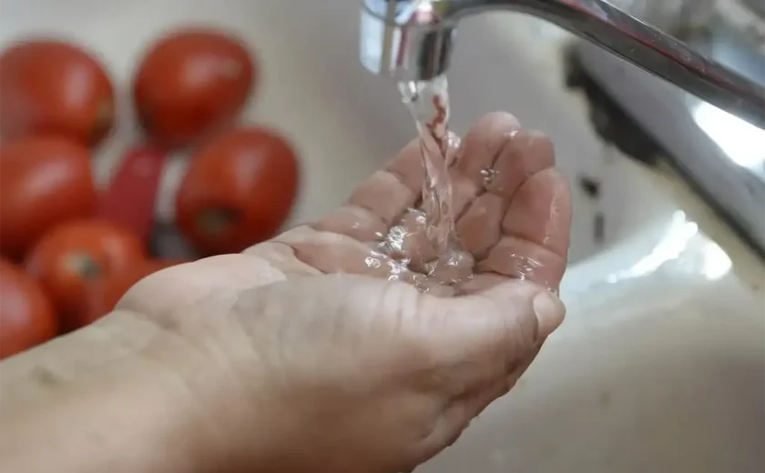 a person's hand positioned under a running tap, with clear water flowing over the fingers. The scene captures the simplicity and freshness of the water, emphasizing cleanliness and daily routine