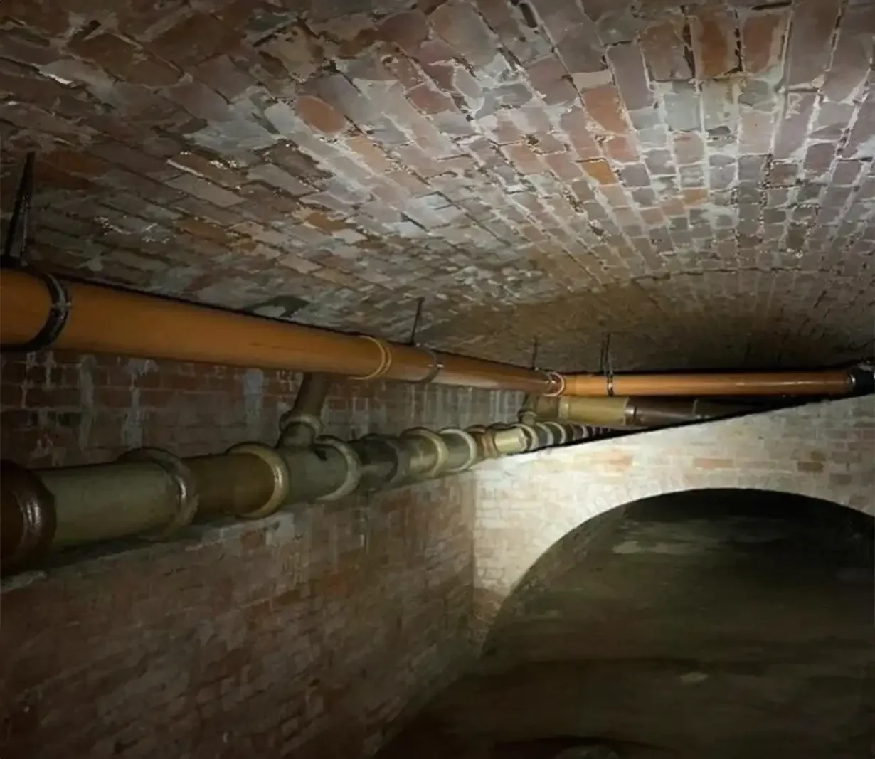 nderground basement featuring a drainage pipe running along the ceiling, with sections made of both cast iron and plastic, securely attached to the ceiling and walls by metal brackets. The setting highlights the infrastructure of the basement, showcasing the integration of various materials in the drainage system
